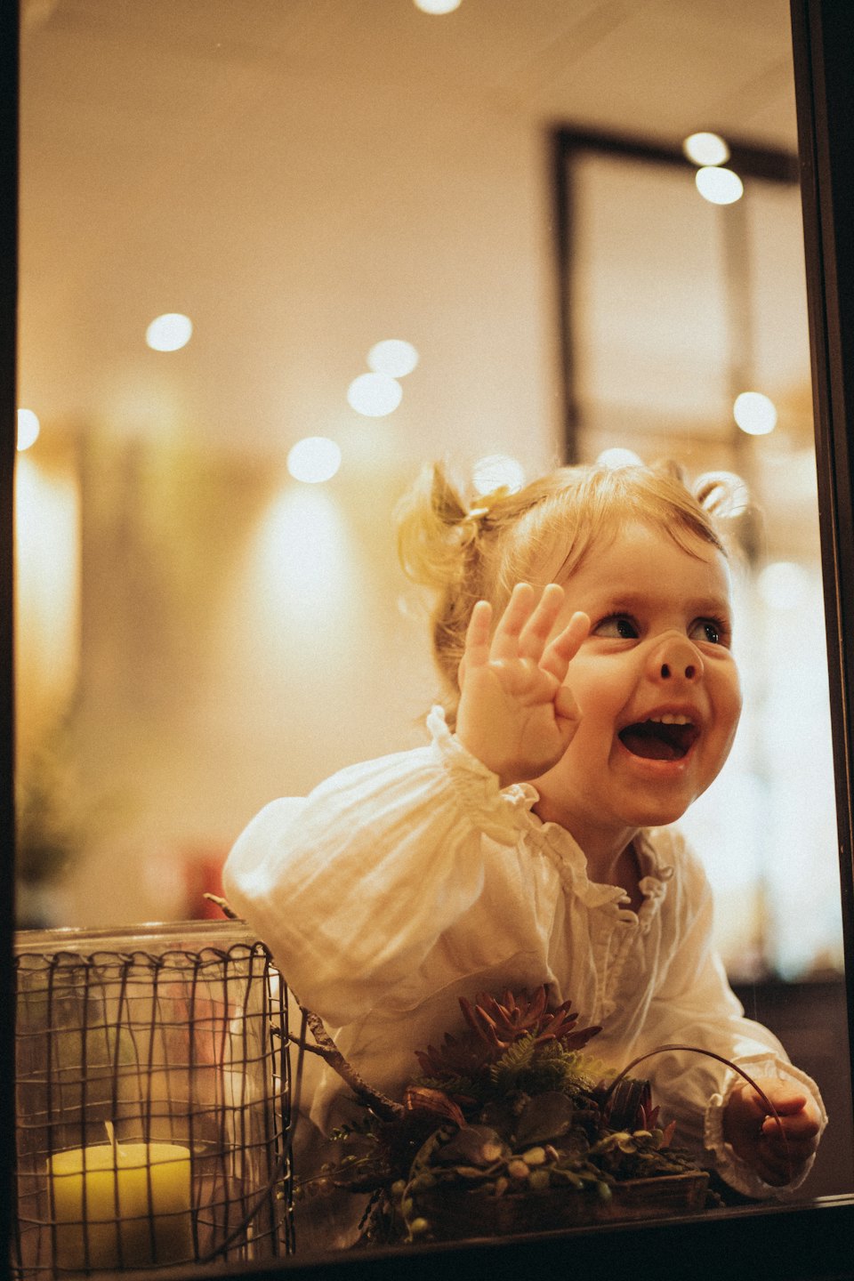 A smiling girl in pigtails waves through a store window.